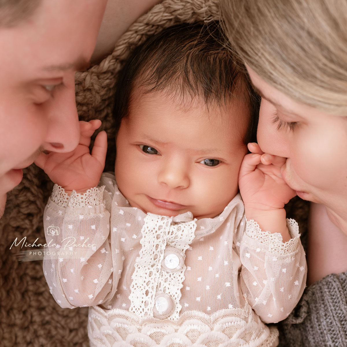 Wachendes Neugeborenes liegt zwischen seinen Eltern, während die Mutter das Baby zärtlich auf die Stirn küsst und der Vater lächelnd nah beim Kind ist.
